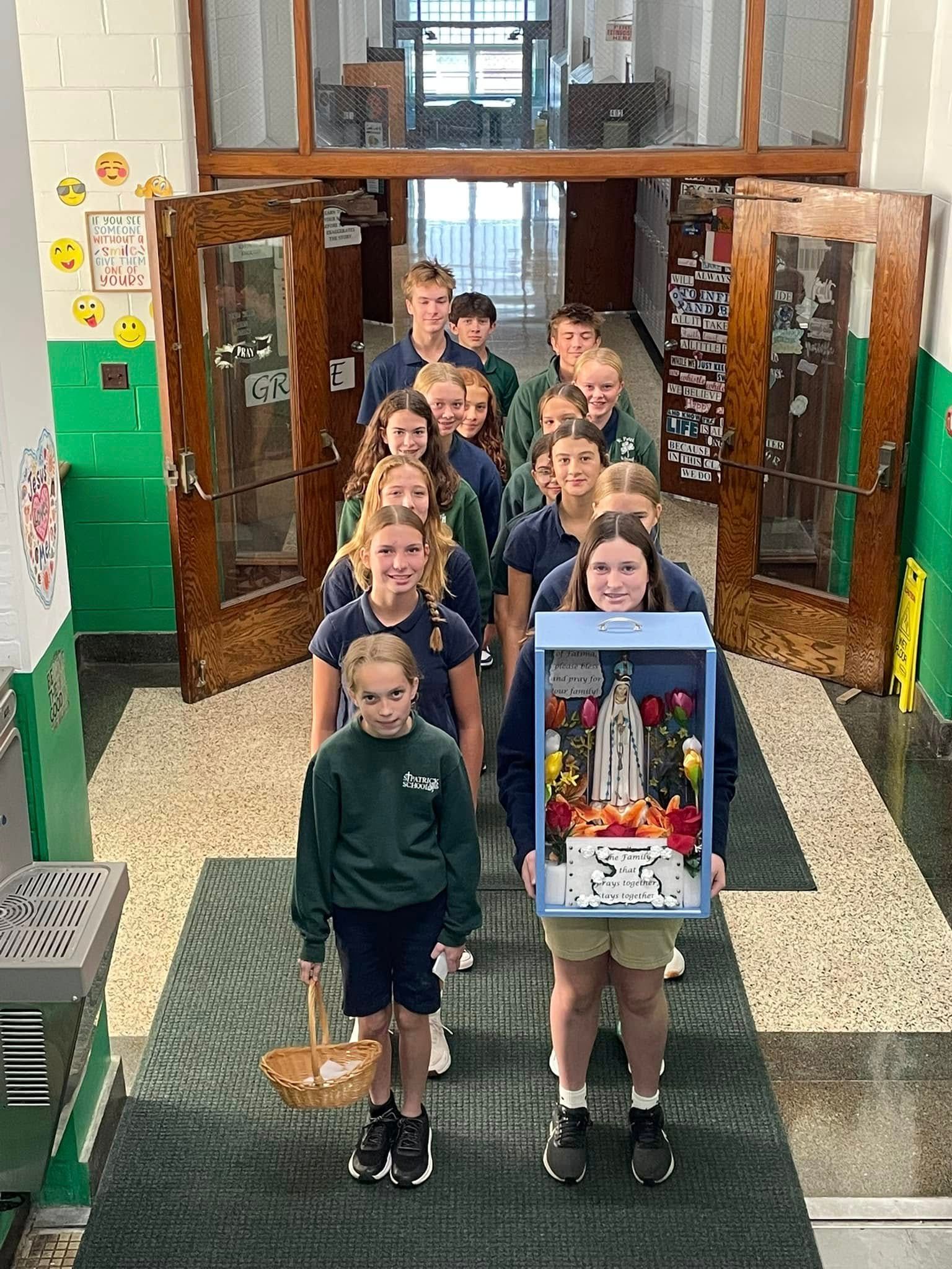 Students in uniforms carry a decorated display and a basket down a school hallway.