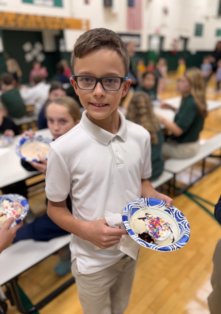 Boy with glasses holds a decorated cookie in a cafeteria, smiling at the camera.