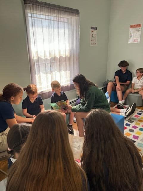 Group of children and young adults listening to a person reading a book in a room with a window.