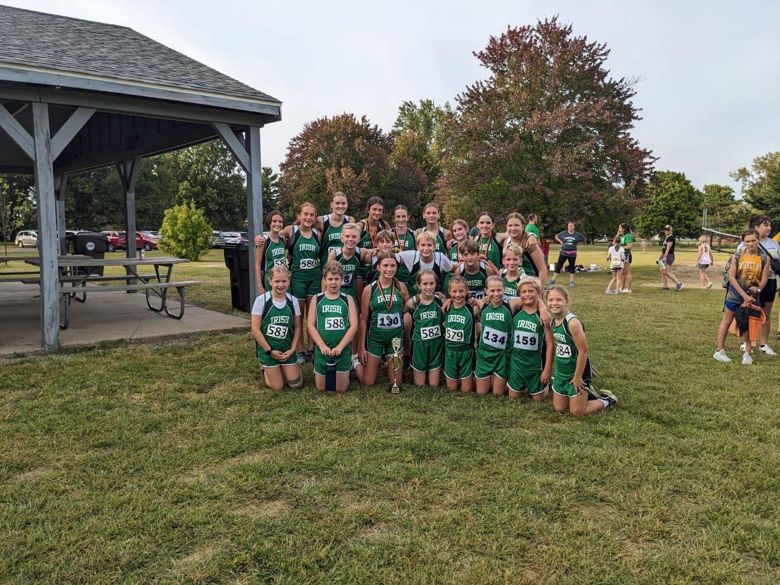 Cross-country team in green uniforms kneels on grass under a gazebo.