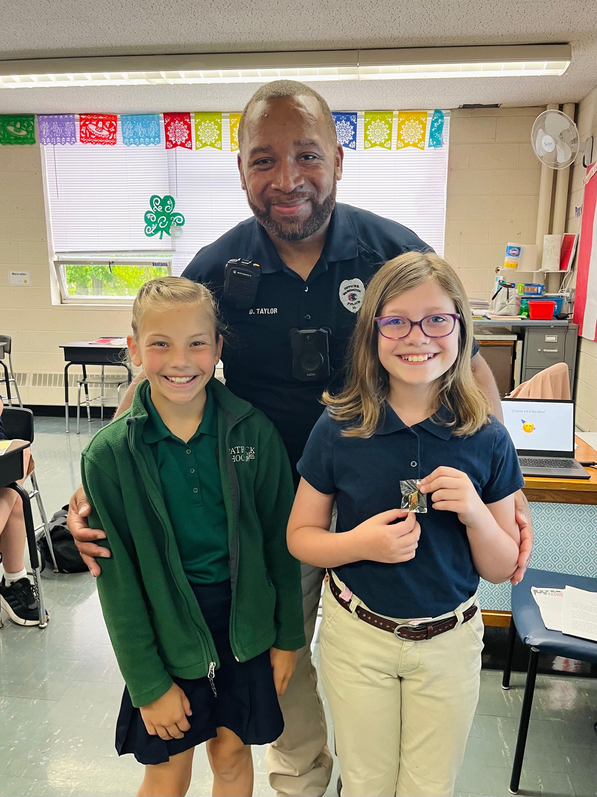 Officer with two smiling girls, one holding a medal, in a classroom.