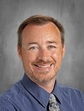 Man with short brown hair and stubble smiles, wearing a light blue and green plaid shirt and green tie.