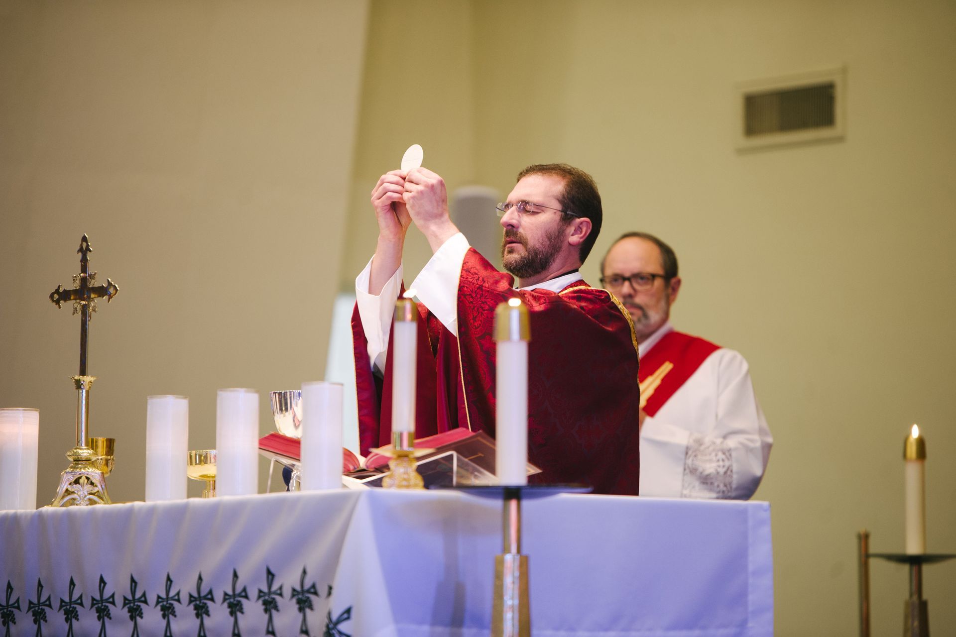 Priest holding up a wafer during Mass, with an altar and attendant, interior of a church.