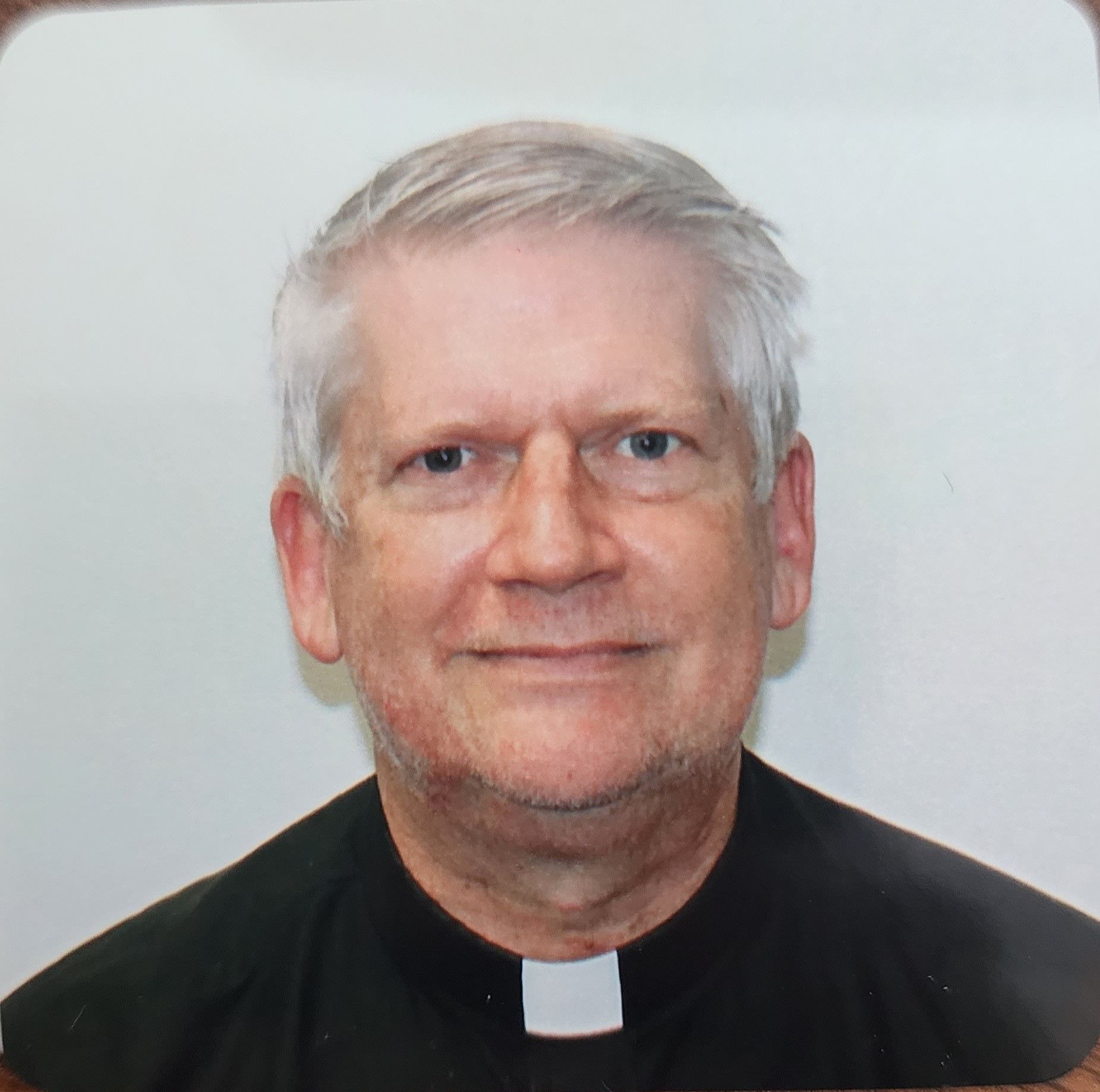 Elderly man with gray hair and clerical collar smiles at the camera.