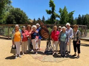 Group of women smiling next to a butterfly sculpture outdoors on a sunny day.