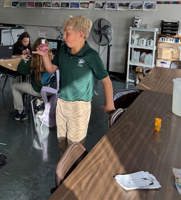 Boy in green shirt, beige shorts, holding object in classroom. Students at desks.