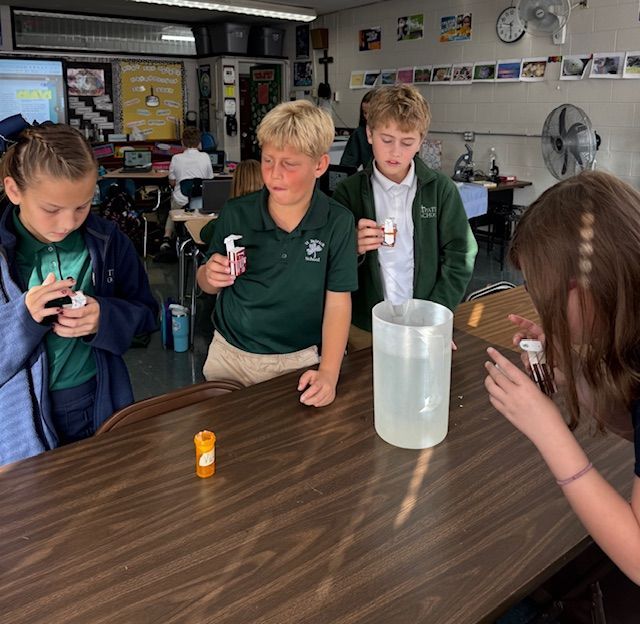 Students in green shirts conduct a science experiment. One looks surprised while others hold vials near a water pitcher.