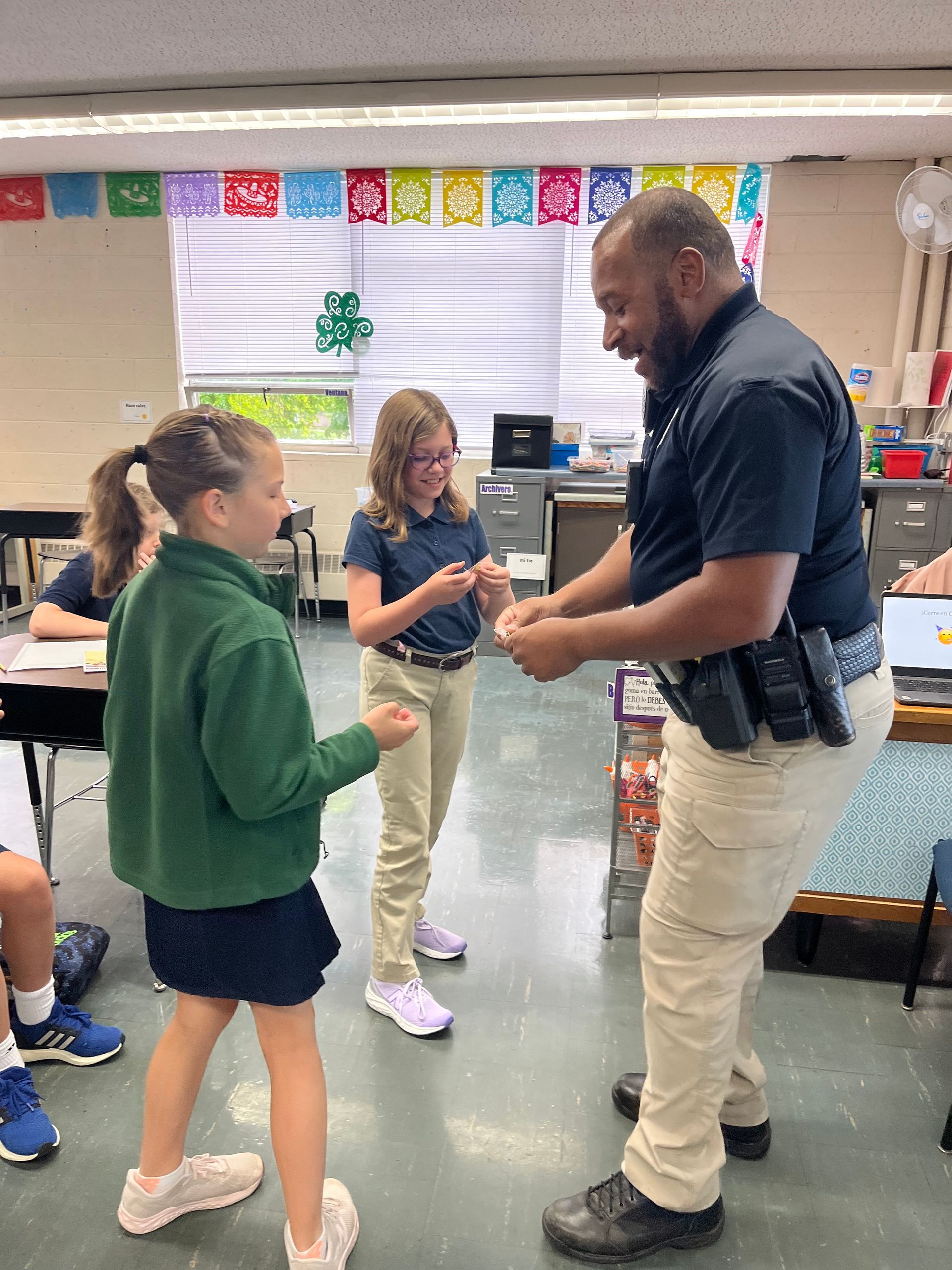 Officer gives a small gift to two smiling students in a classroom.