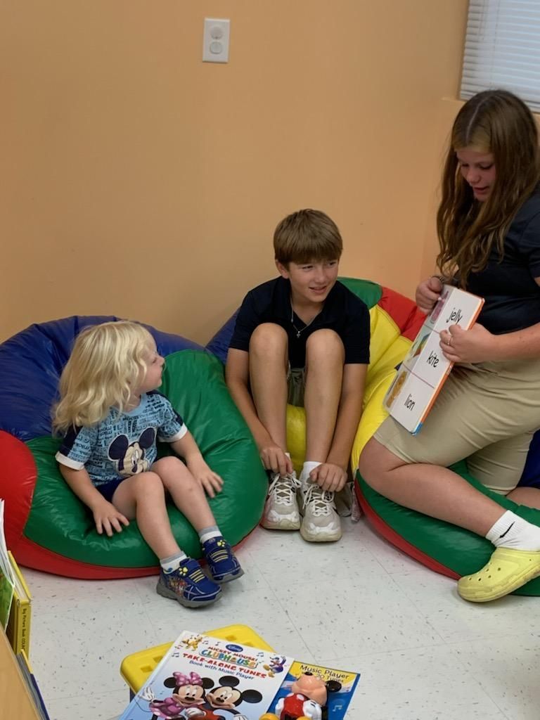 Three children in a room. One reads from a book, the other two listen on bean bags.