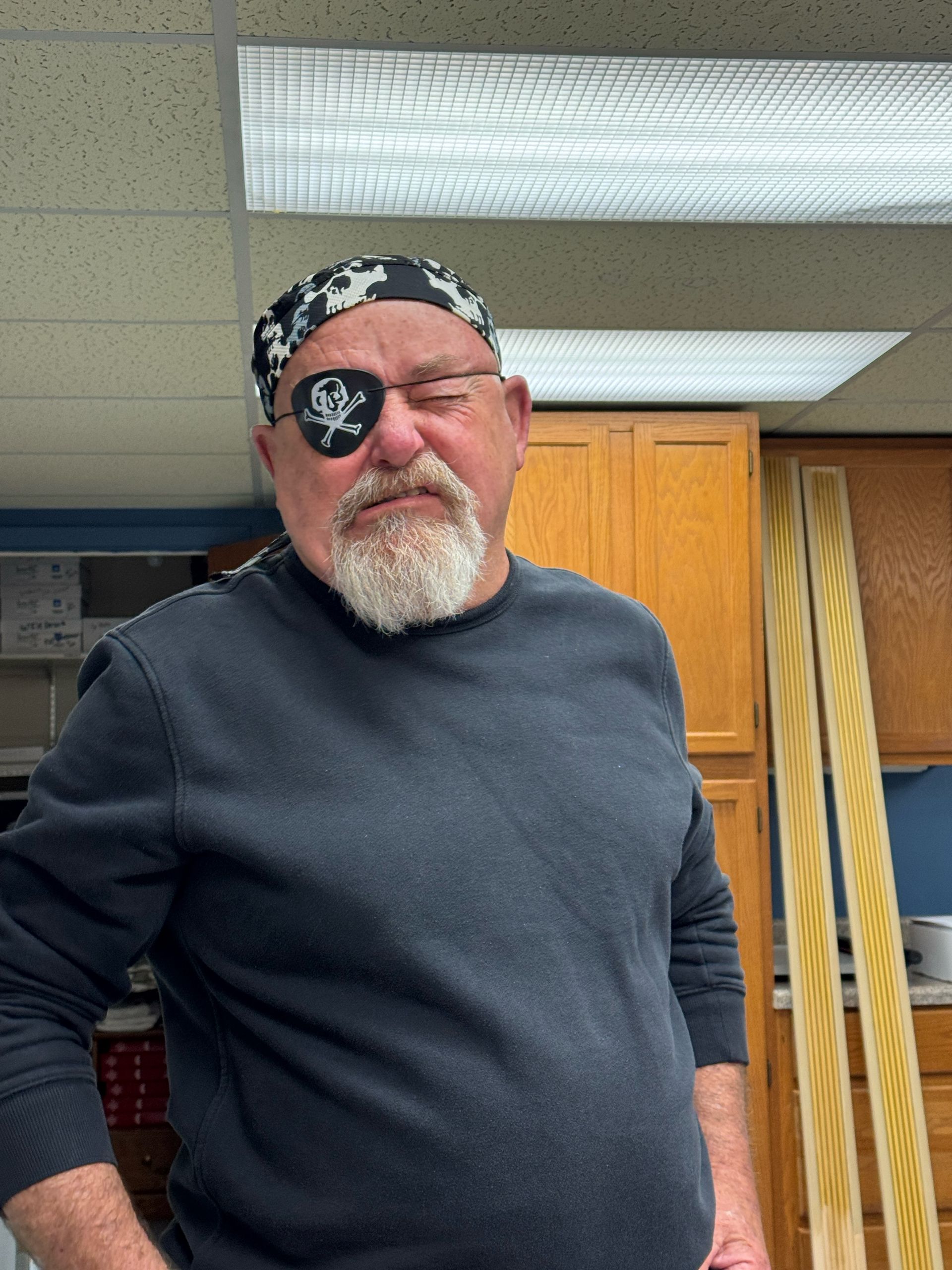 Man wearing pirate eye patch, bandana, and gray beard. Leaning against a wooden cabinet.