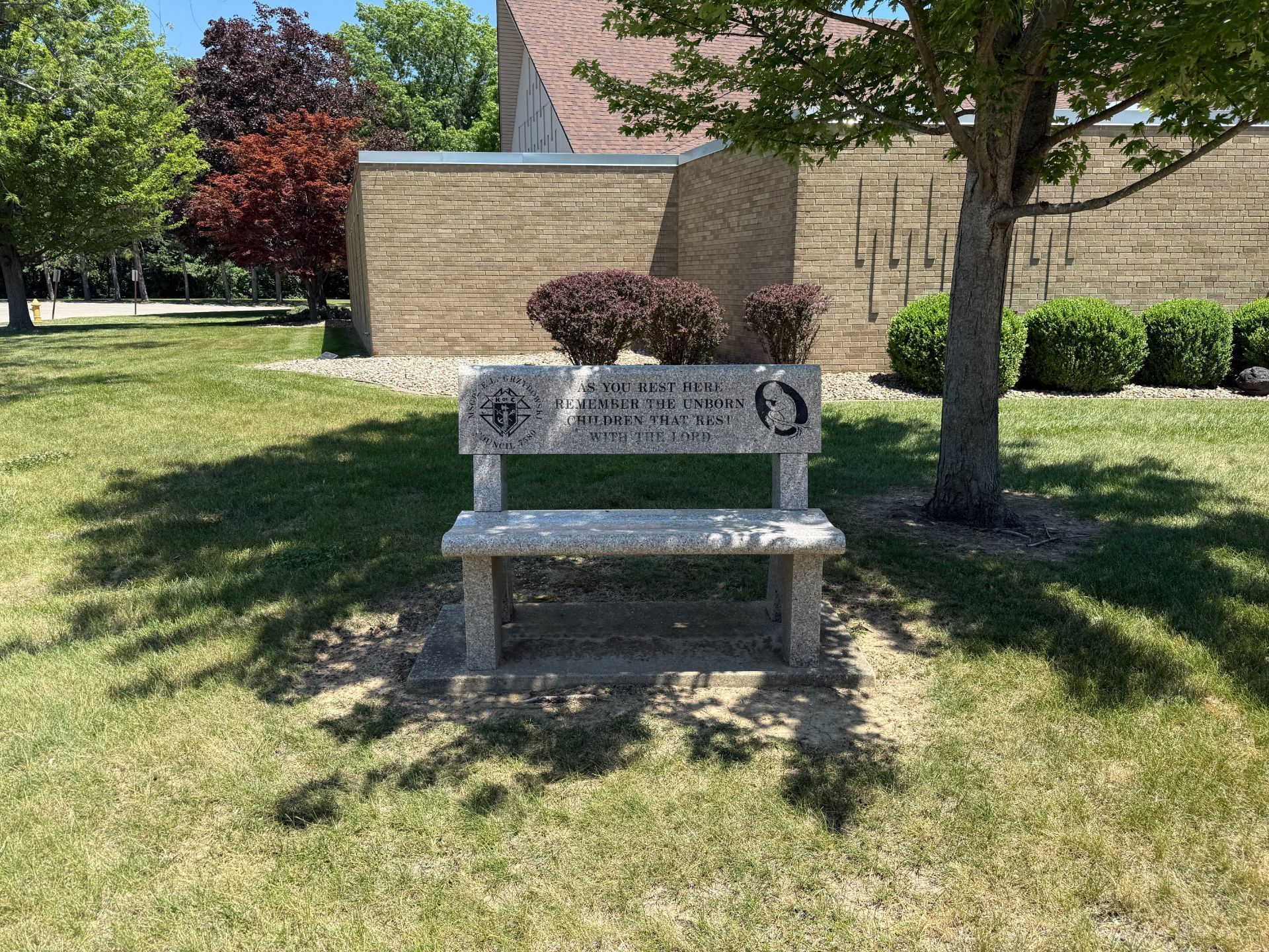 A stone bench under a tree in a grassy area. Behind the bench is a building with a brick facade.