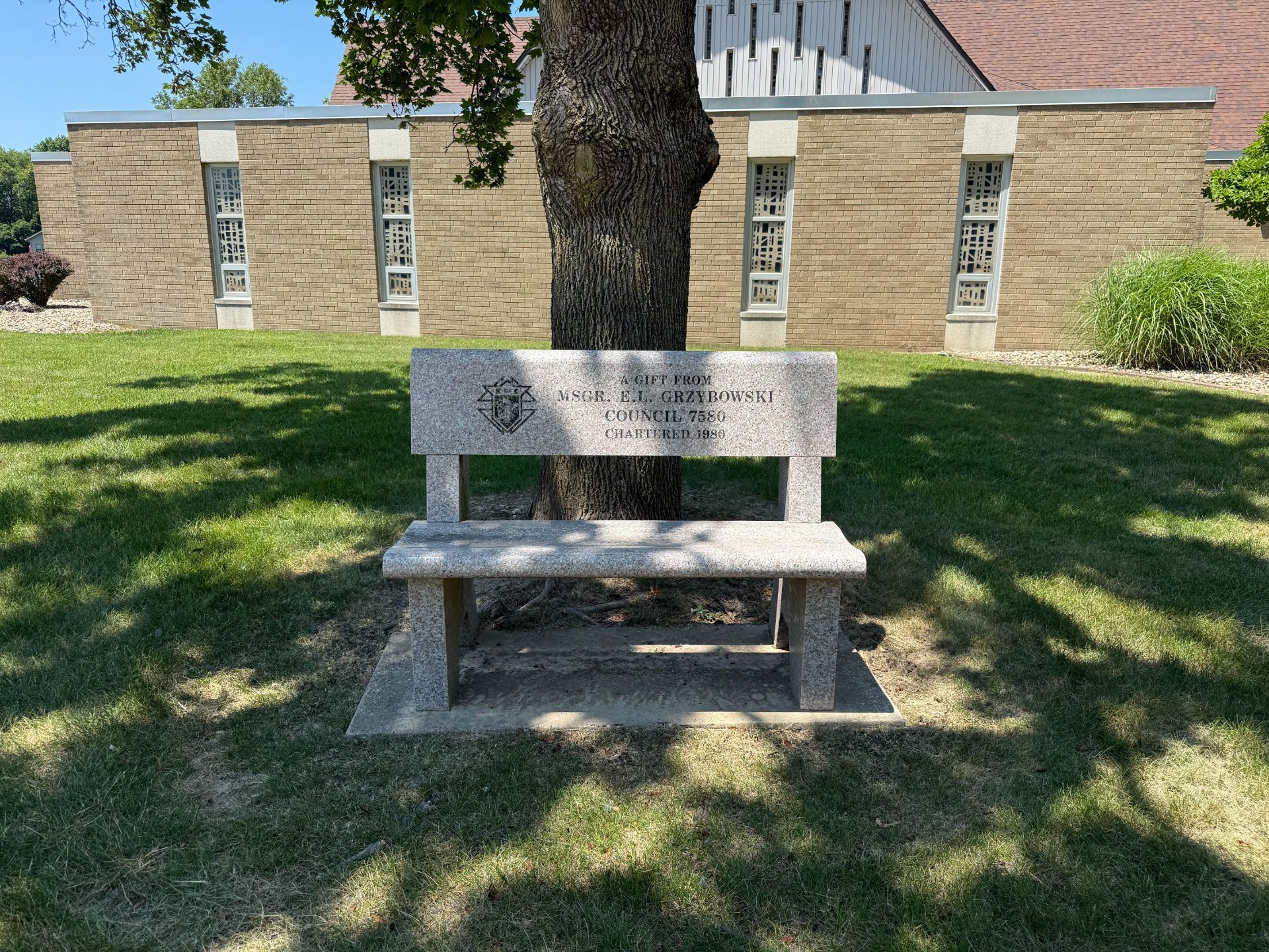 Granite bench with inscription under a tree in front of a church.