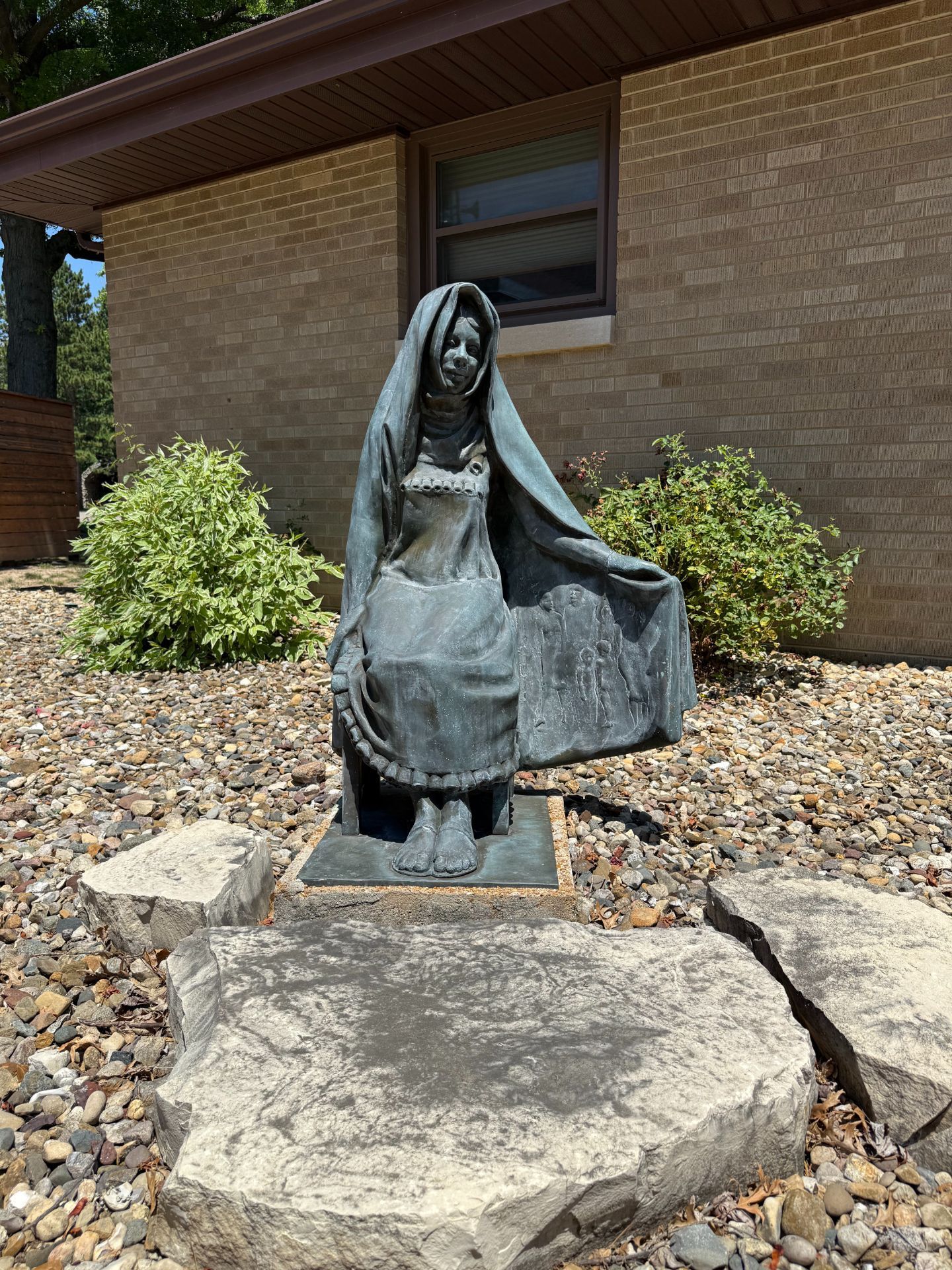 Bronze statue of woman seated, wearing a veil, outdoors in front of a brick building.