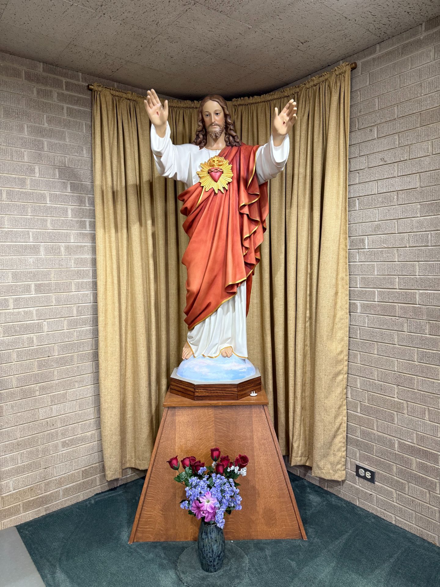 Statue of Jesus with open arms, red robe, heart exposed, on a pedestal with flowers, against gold curtains, brick walls.