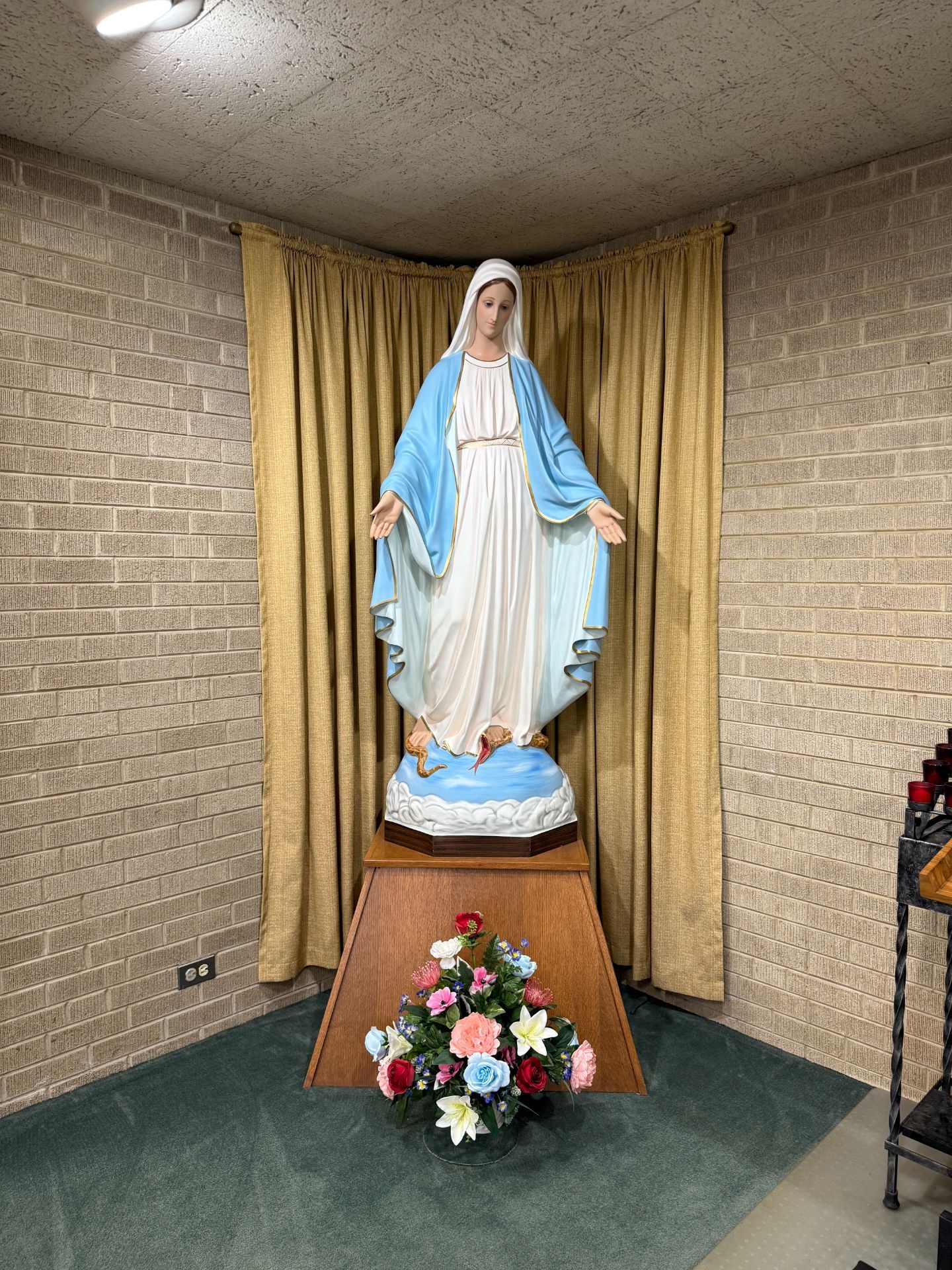 Statue of Virgin Mary in a corner alcove, hands outstretched, wearing blue and white robes. Flower arrangement in front.