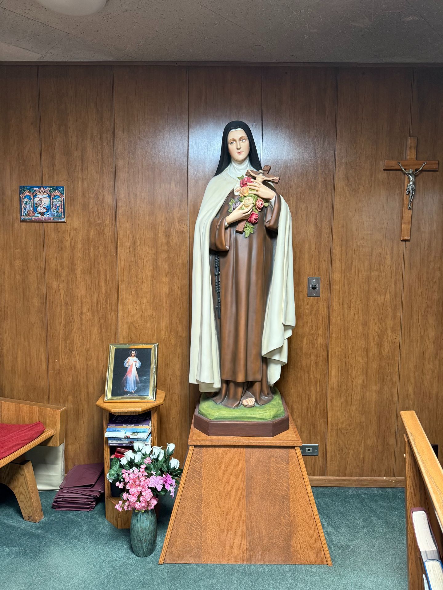 Statue of a saint holding flowers, wooden paneling background, cross on the wall.