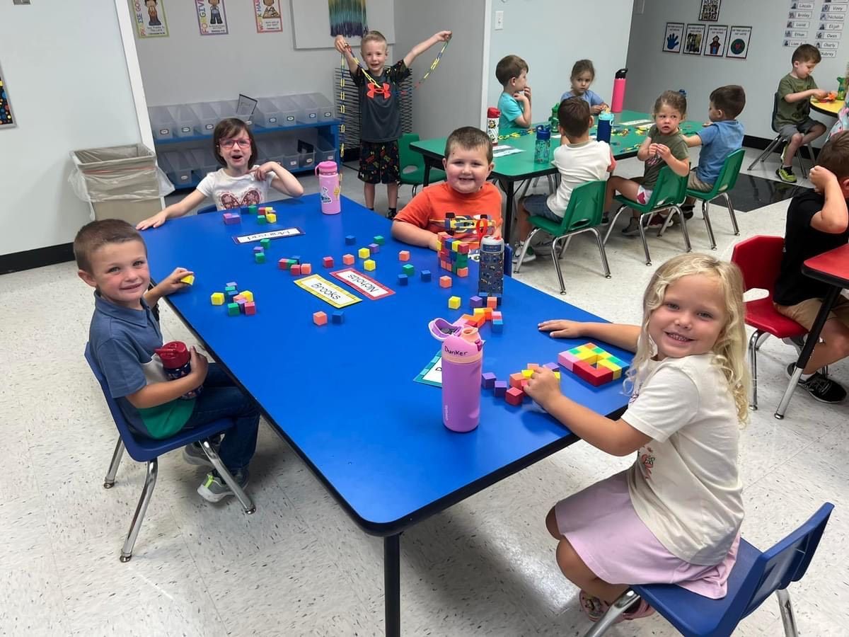 Children at a blue table playing with blocks in a brightly lit classroom.