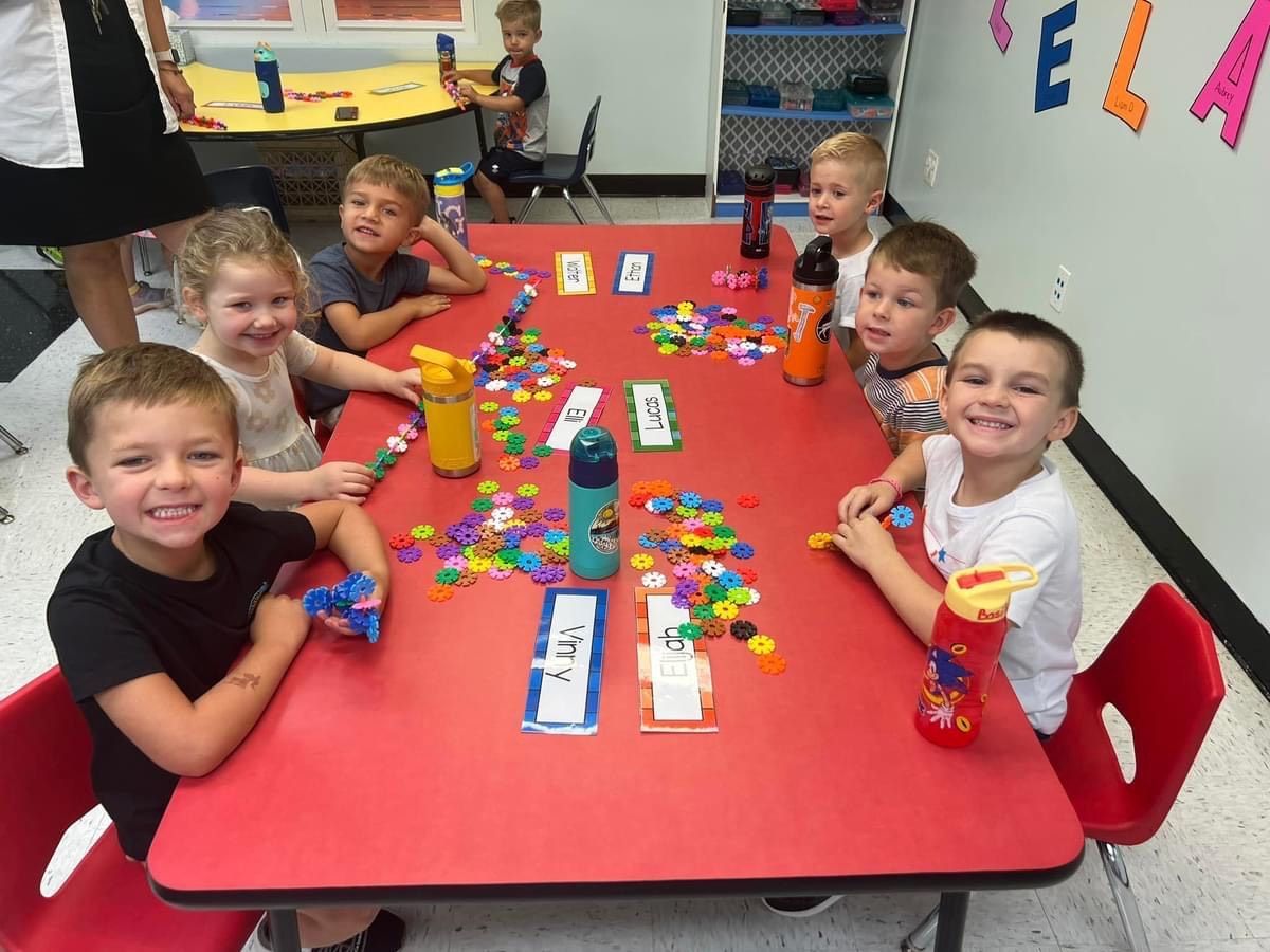Children at a red table smiling, playing with toys and looking at the camera in a classroom.