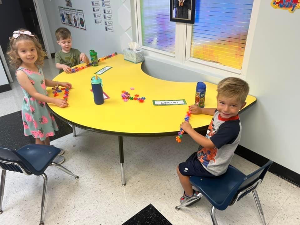 Three young children playing with toys at a yellow table in a classroom.