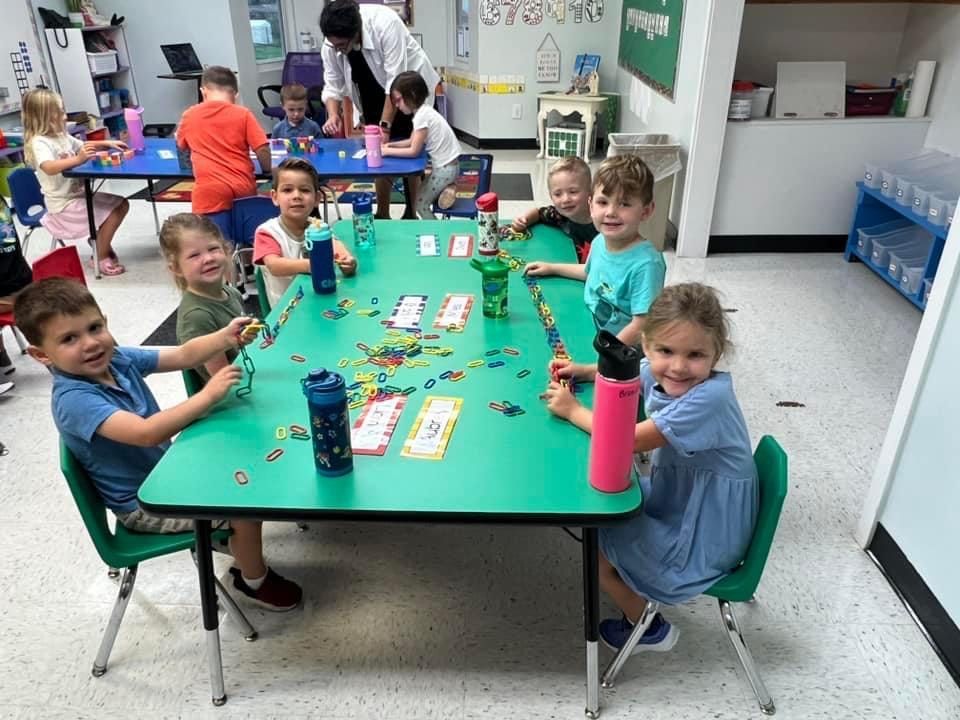 Children sitting around a green table, playing with colorful toys, teacher in the background.