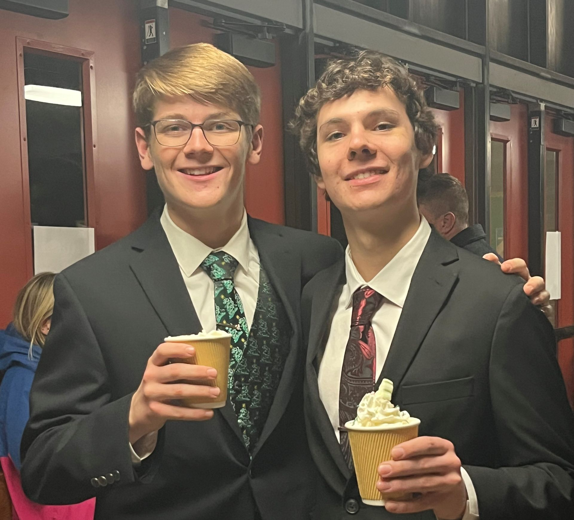 Two young men in suits smile, holding coffee cups with whipped cream. Inside a building with dark red walls.