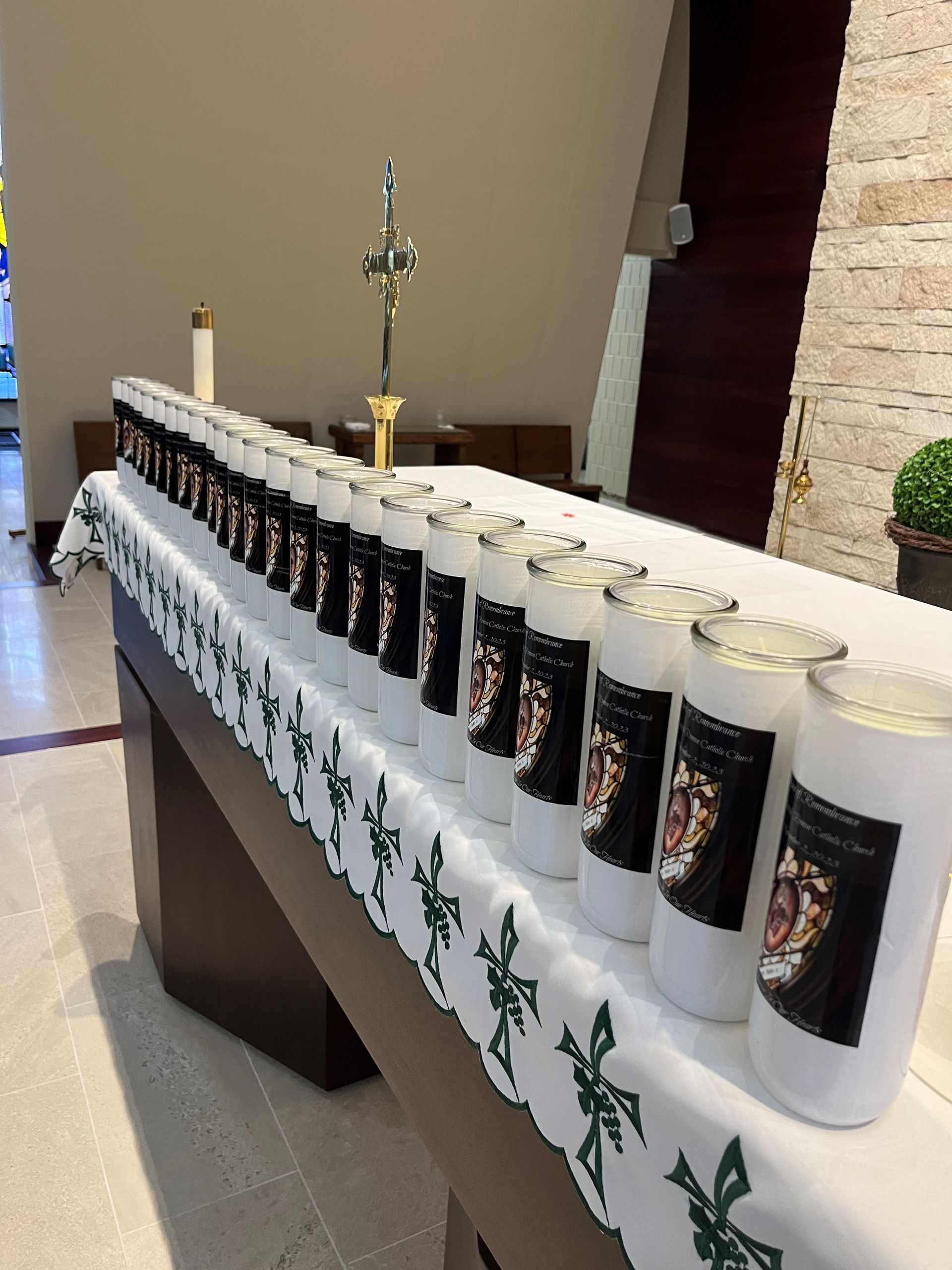 Candles lined on a table with a cross in a church setting. White table cloth with green cross design.