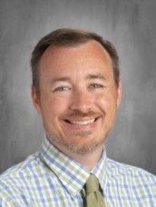 Man with short brown hair and stubble smiles, wearing a light blue and green plaid shirt and green tie.