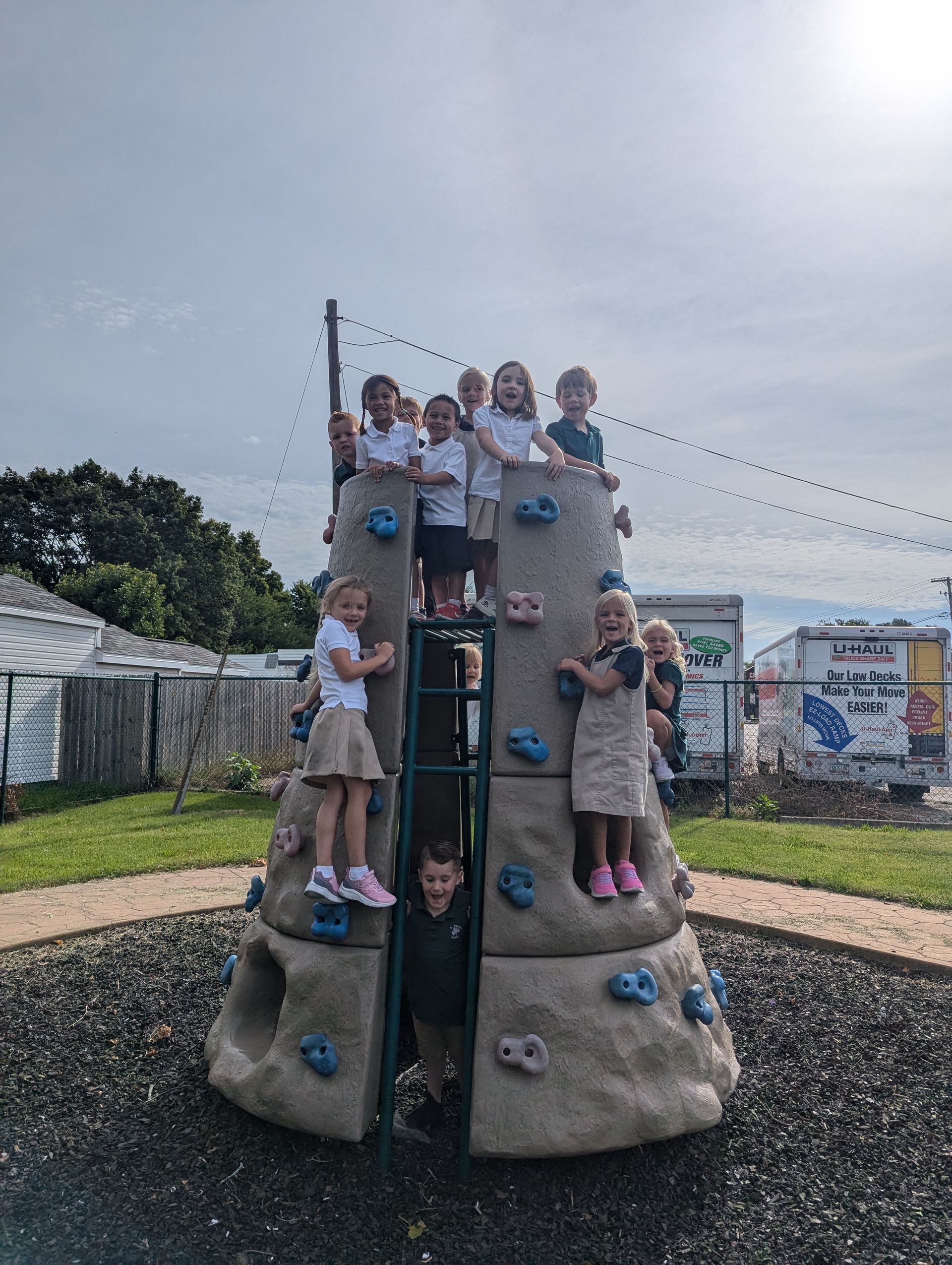 Children playing on a gray, cone-shaped climbing structure with blue handholds at a playground.