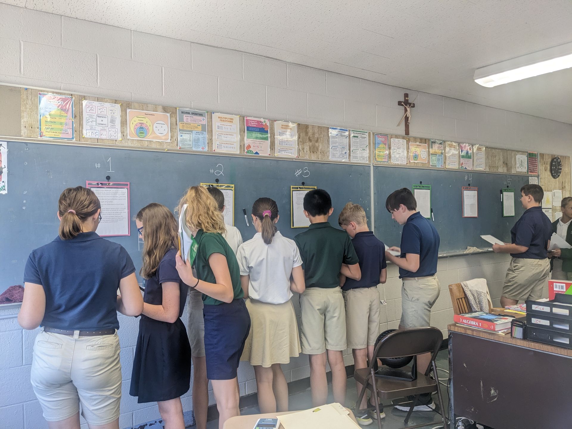 Students in uniform at a chalkboard, examining papers and projects in a classroom.