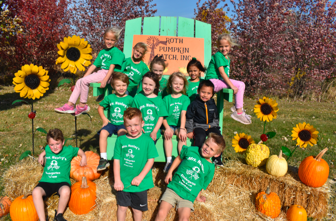 Group of children in green shirts at a pumpkin patch, posing by sunflowers and hay bales.