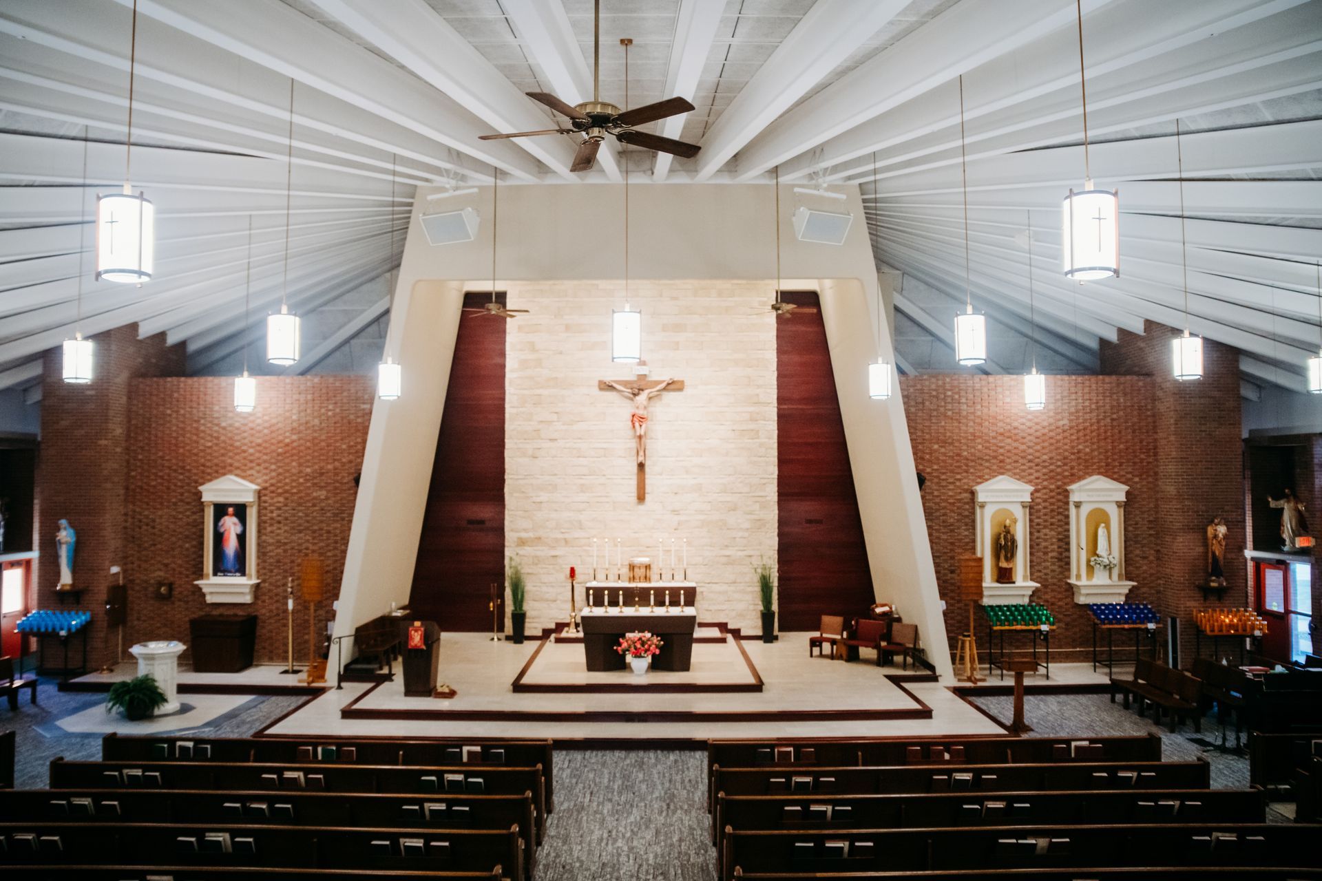 Interior view of a church with altar and crucifix, brick walls, rows of pews, and hanging lights.