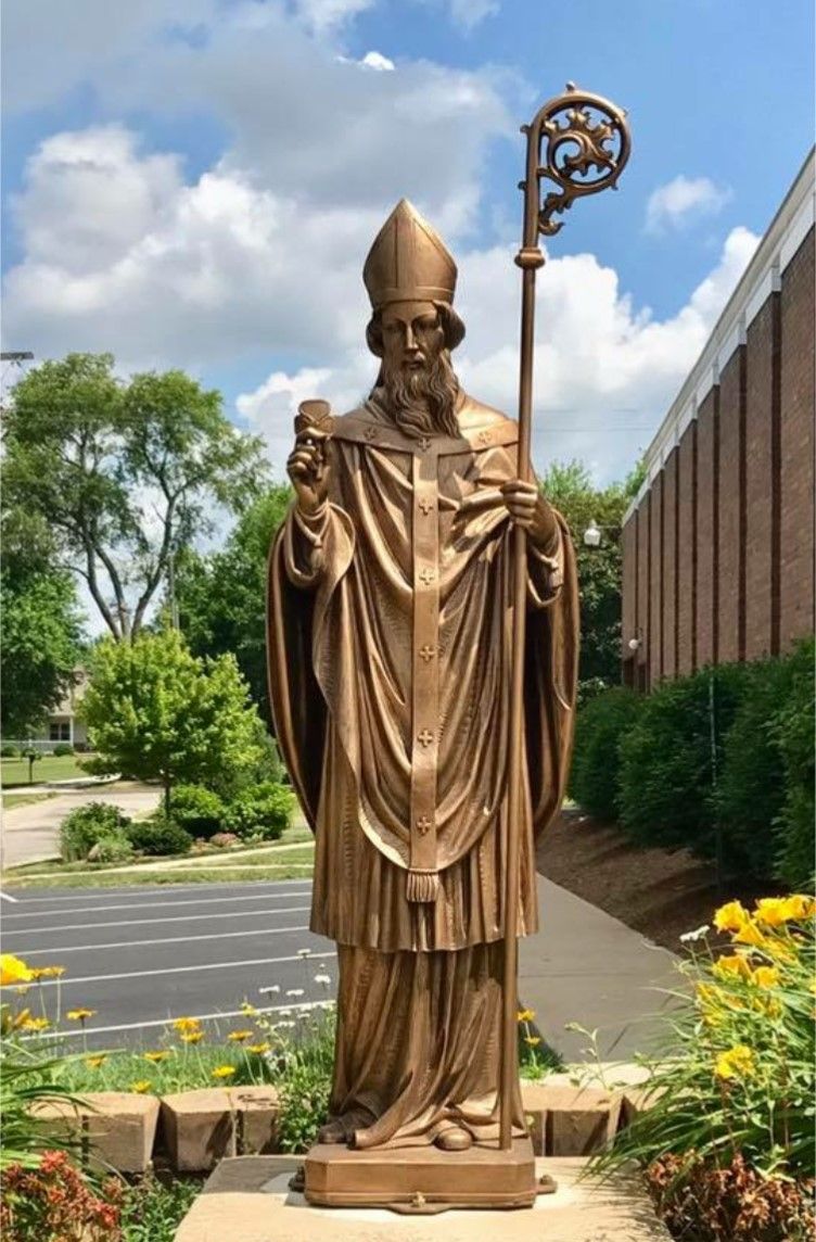 Bronze statue of a bishop holding a staff and cross, standing outside a building.