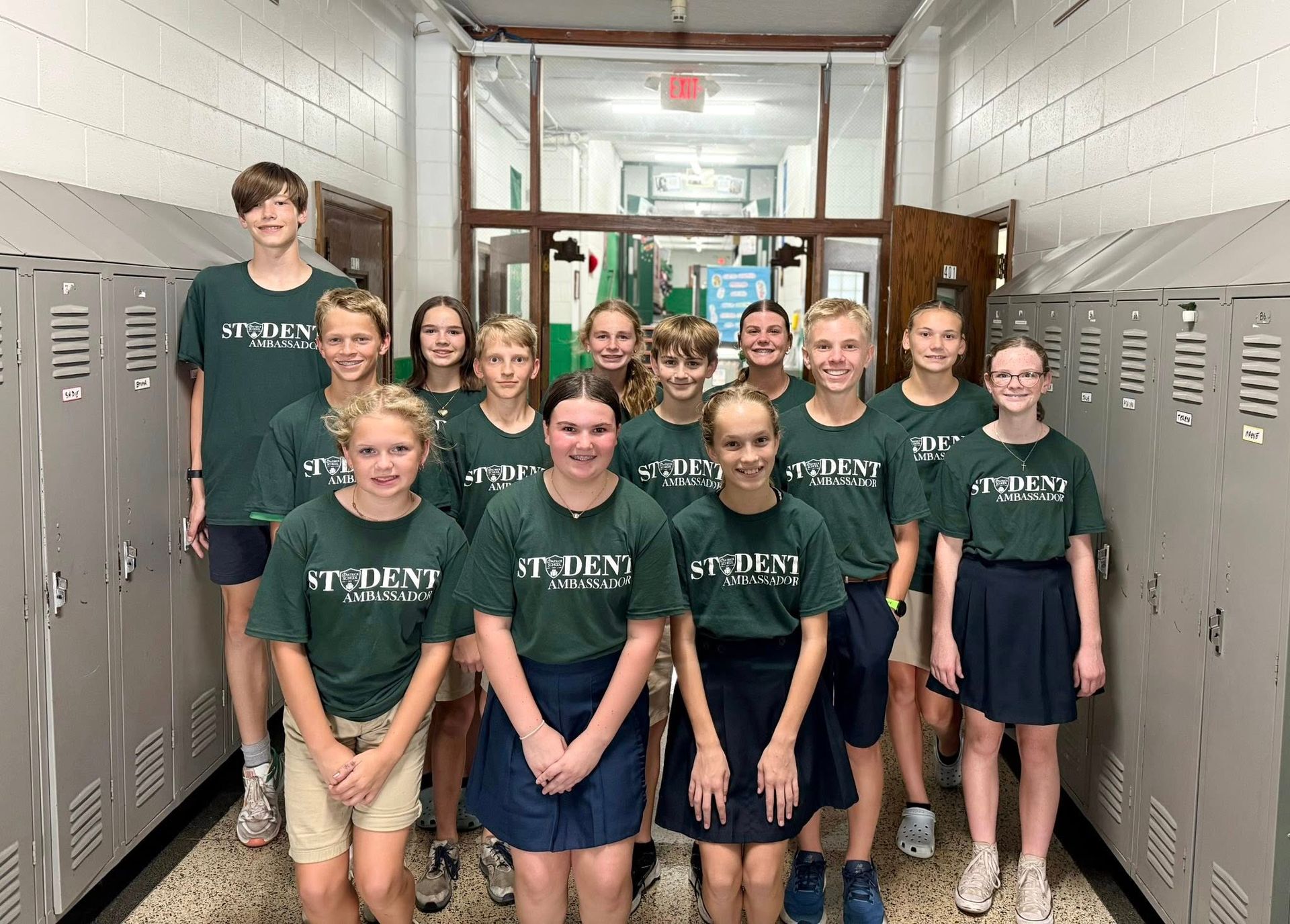 Group of students in green shirts and skirts/shorts in a school hallway, smiling.