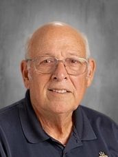 Older man with glasses, smiling in a navy shirt, set against a gray backdrop.