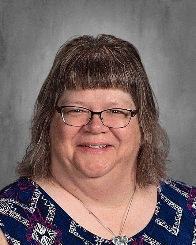 Woman with short brown hair and glasses smiles, wearing a patterned blue top.