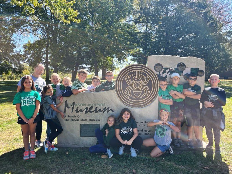 Children pose at the Nelson Atkins Museum sign. They are wearing green shirts and smiling on a sunny day.