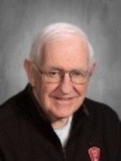 Smiling elderly man with white hair and glasses, wearing a black jacket.