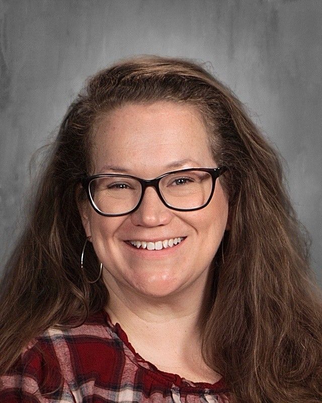 Woman with glasses smiles, wearing a red plaid shirt, long brown hair.