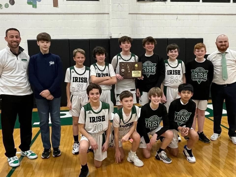 A youth basketball team in green and white uniforms poses with a trophy in a gymnasium.