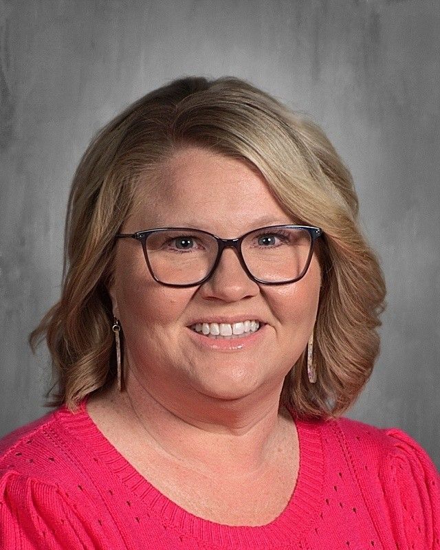 Woman with blonde hair and glasses, wearing a pink sweater, smiling.