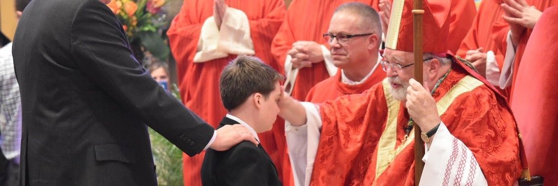 A priest anoints a boy's forehead during a religious ceremony. Men in red robes surround them.