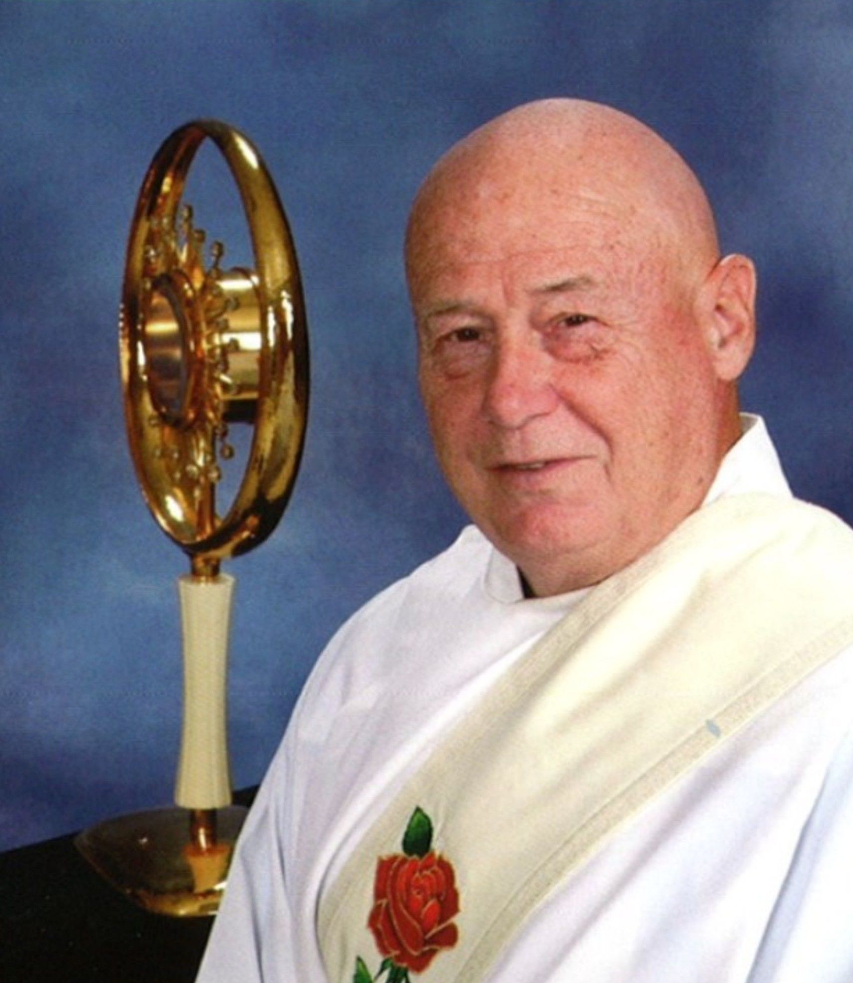 Bald priest in white vestments smiles, with a golden monstrance to his left.