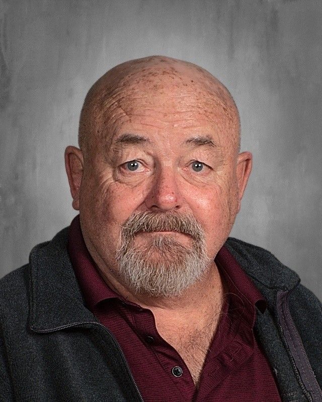 Bald, older man with a gray beard and blue eyes. He wears a maroon shirt and a dark jacket, looking directly at the viewer.