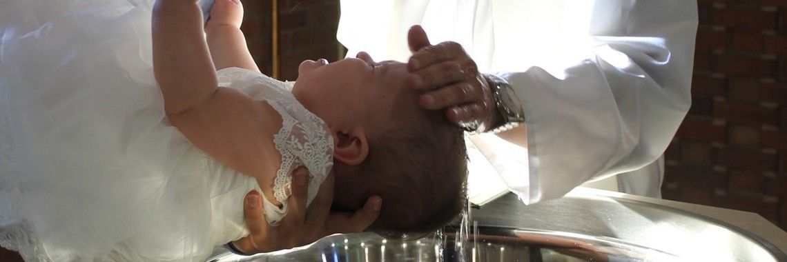 A baby is baptized in a metal basin by a person in a white robe.