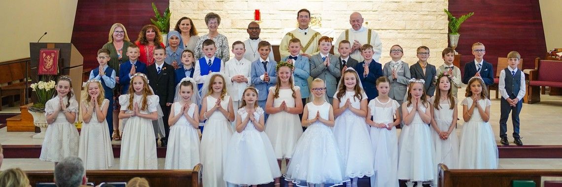 Group of children in white dresses and suits, with clergy, in a church setting. Praying, solemn expressions.