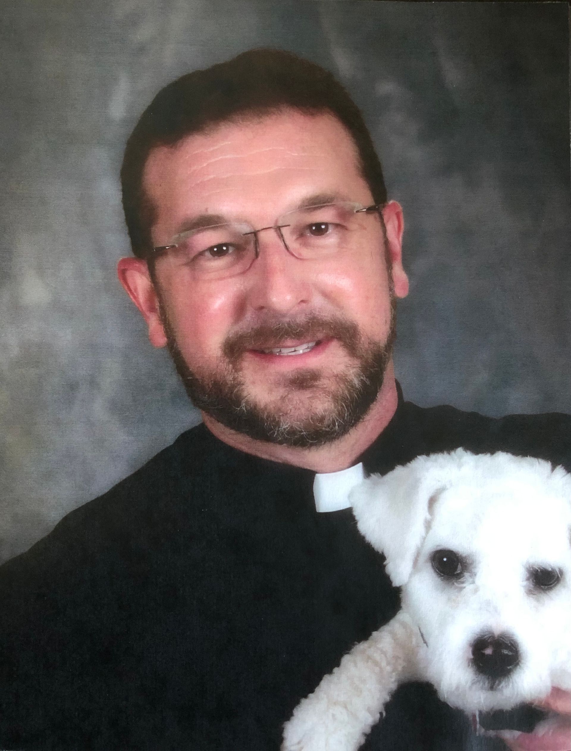Man in clerical collar holding a small white dog, smiling.