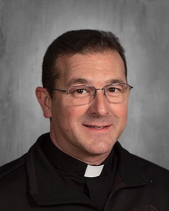Priest with glasses and clerical collar smiling, against a gray backdrop.