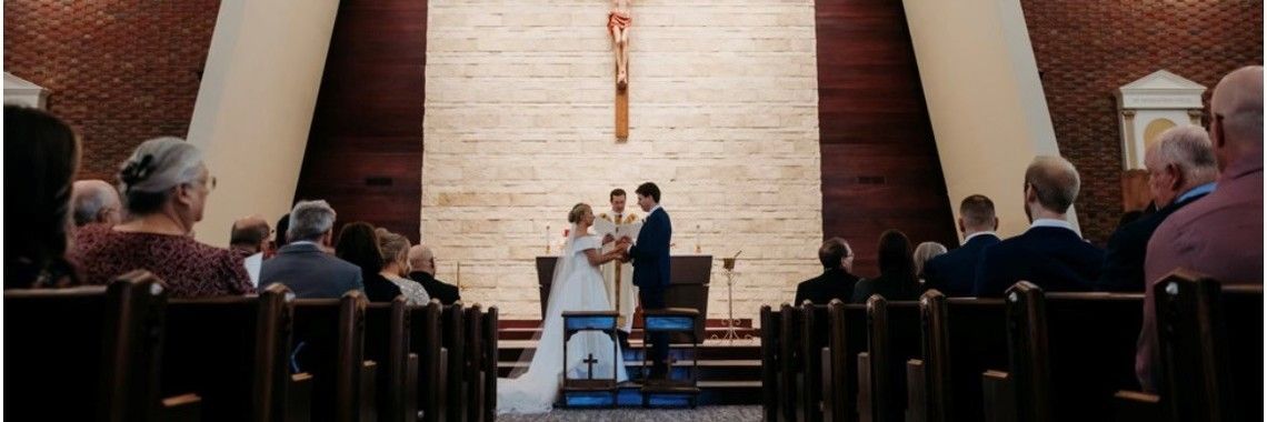 Wedding ceremony in a church, bride and groom exchanging vows, attendees seated. Crucifix on the wall.