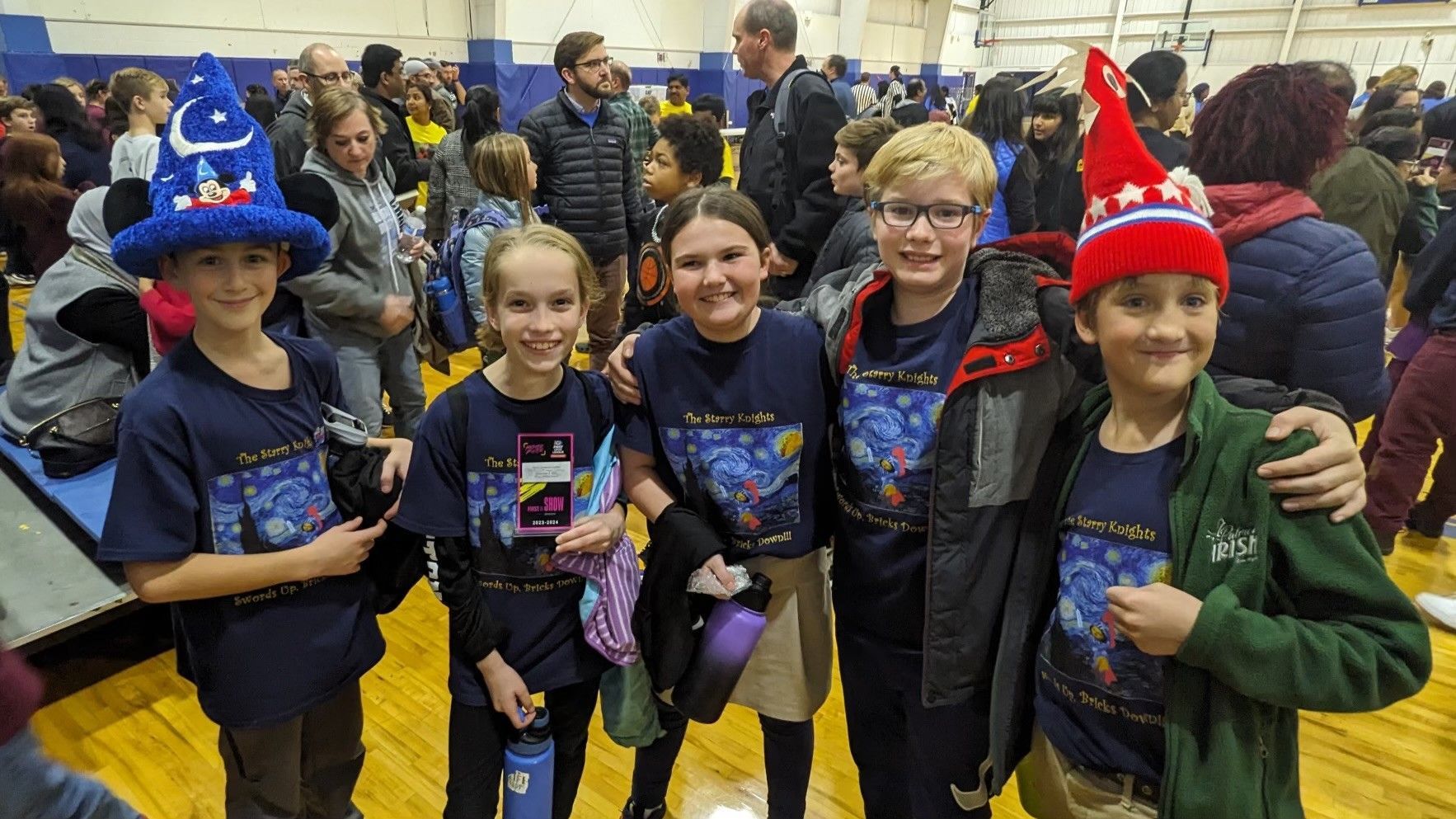 Group of children in blue shirts and hats posing for a photo in a gym.