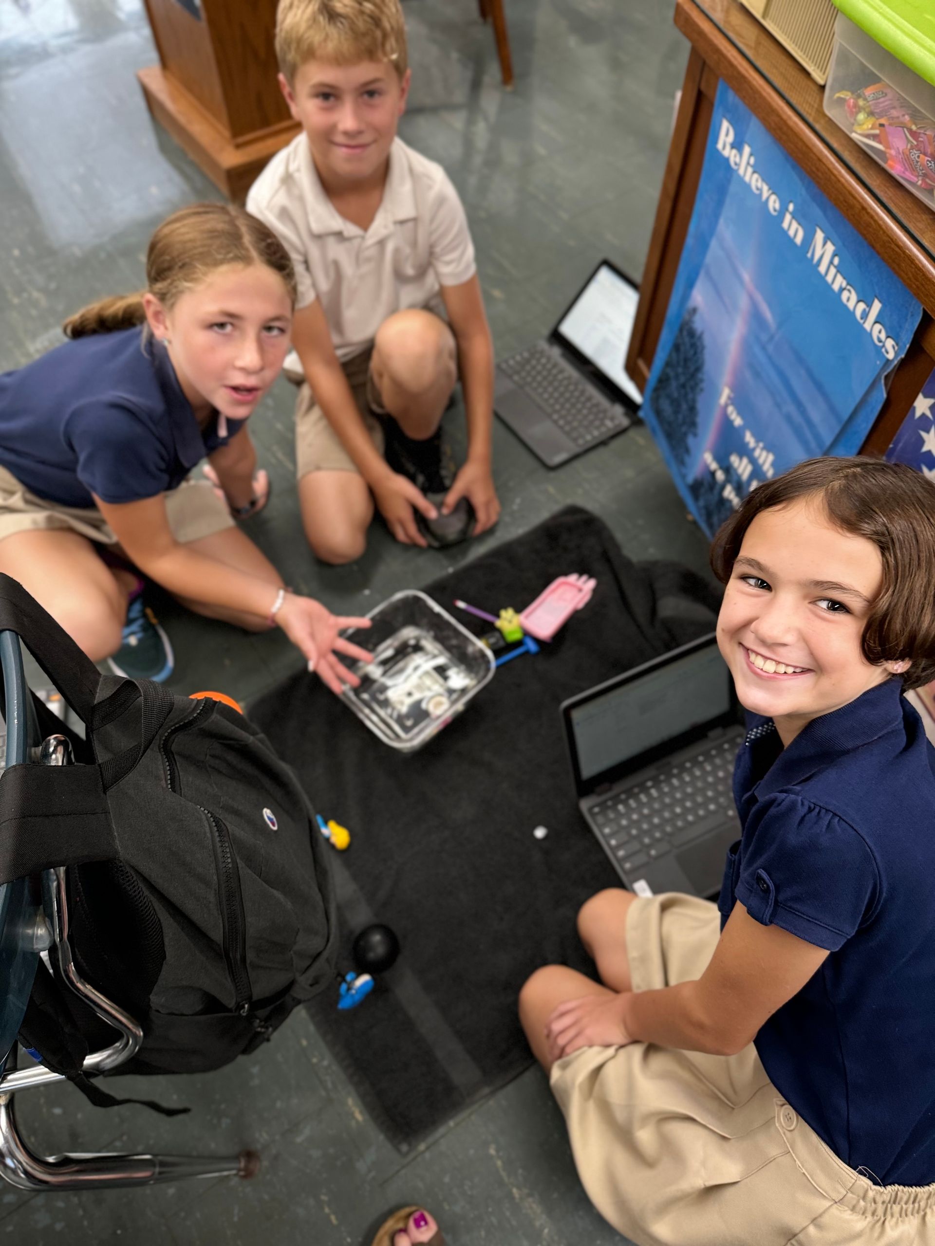 Three students sit around a black mat, with laptops and a container. They look up, smiling.